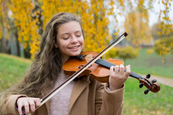 Girl playing the violin in the autumn park at a yellow foliage background. Young violinist playing in the park