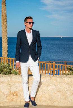 Takım elbiseli genç yakışıklı bir adam ve deniz dibinde beyaz pantolon ve palmiye ağaçları. Moda Modeli