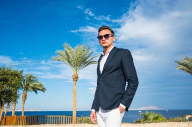 Takım elbiseli genç yakışıklı bir adam ve deniz dibinde beyaz pantolon ve palmiye ağaçları. Moda Modeli