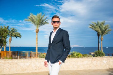 Takım elbiseli genç yakışıklı bir adam ve deniz dibinde beyaz pantolon ve palmiye ağaçları. Moda Modeli