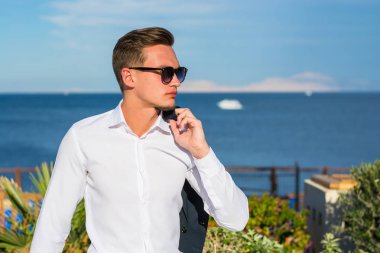 Bir takım elbise ve beyaz pantolon deniz arka plan ve palmiye ağacının genç yakışıklı adam. Moda Model