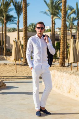 Beyaz gömlekli, beyaz pantolonlu, palmiye ağaçlarının arkasında plajda oturan yakışıklı bir adam. Moda Modeli