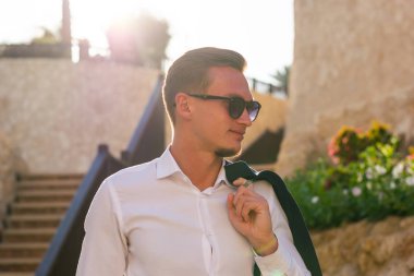 Genç adam beyaz bir gömlek ve güneş gözlüğü standları ve yakınındaki gülümsüyor