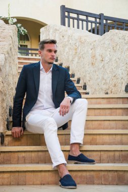 Genç adam bir takım elbise ve beyaz pantolon içinde merdivenlerde oturur