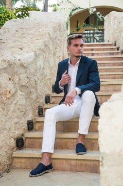 Genç yakışıklı olan bir takım elbise ve beyaz pantolon STAI üzerinde oturur