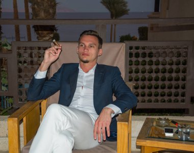 Takım elbiseli genç yakışıklı bir balcon üzerinde oturan bir puro içiyor