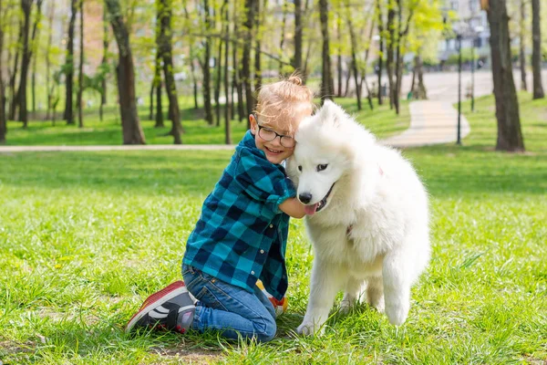 Маленький хипстерский мальчик в очках обнимает белую пушистую самодовольную собаку — стоковое фото