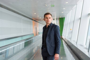 Takım elbiseli genç bir adam yürüyen merdivenin yanında havaalanında duruyor.