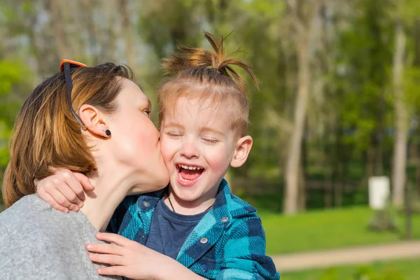 Мама целует своего мальчика, который смеется в парке — стоковое фото