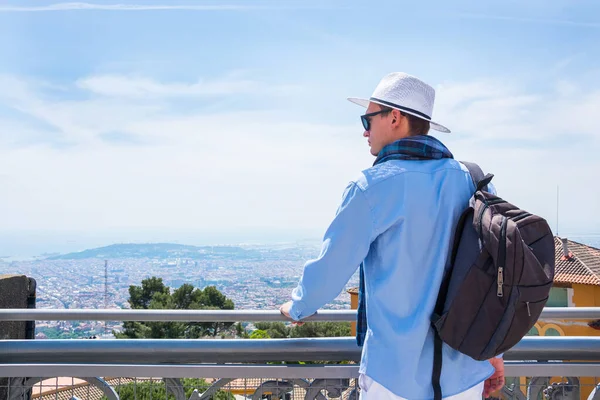 A young man in a scarf, in hat and sunglasses with a backpack stands near the Temple of the Sacred Heart on Mount Tibidabo in Barcelona, Spain at the beautyful landscape background