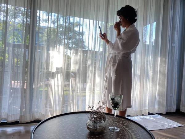 Woman in hotel room or apartment. Girl in white bathrobe stands in front of full-length window with smartphone and cup of coffee.