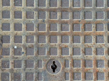 Geometrik kare şablon ve anahtar desenli eski paslı metal rögar kapağı, kentsel dokuyu, endüstriyel tasarımı ve grunge sokak arka plan detaylarını gösteriyor.