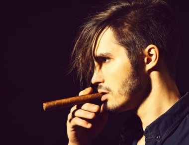 Yakışıklı, sakallı, mavi gömlekli, şık saçlı, ciddi yüzlü, siyah stüdyo arka planında puro içen bir adam.