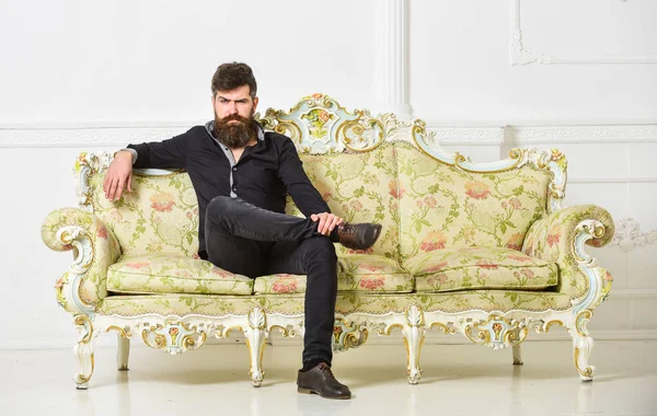 Hipster en cara estricta se sienta solo. Concepto rico y solitario. Propietario de apartamento de lujo se sienta en el sofá, fondo blanco de la pared. Hombre con barba y bigote pasa el ocio en la sala de estar de lujo — Foto de Stock