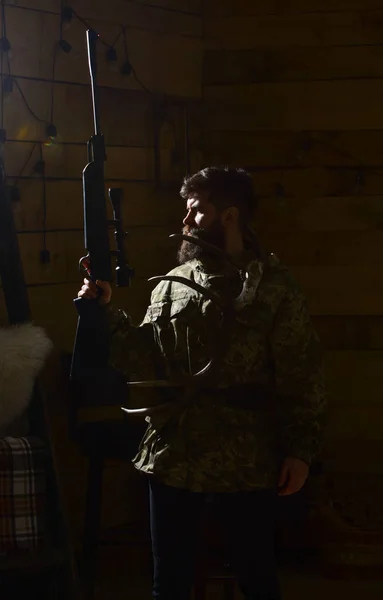 Concepto Hunter. El hombre con barba lleva ropa de camuflaje, fondo oscuro. Macho en cara estricta en la casa de guardabosques listo para la caza. Cazador brutal con arma y cuernos de ciervo, iluminado en la oscuridad —  Fotos de Stock