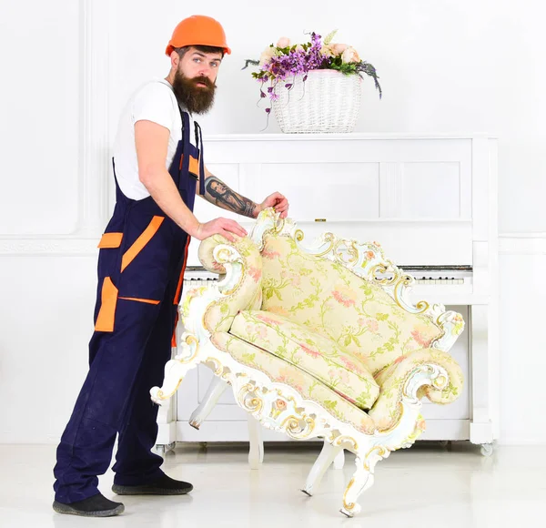 Relocating concept. Courier delivers furniture in case of move out, relocation. Man with beard, worker in overalls and helmet lifts up armchair, white background. Loader moves armchair for move out