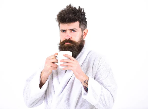 Macho drowsy, sleepy with strict face drinks coffee in morning. Morning rituals concept. Man with beard and disheveled hair stands in bathrobe, holds mug with tea or coffee, white background — Stock Photo, Image
