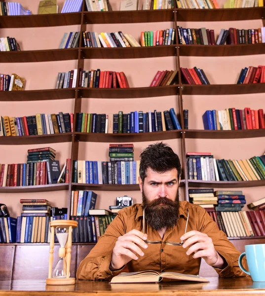 男人在忙碌的脸上看书, 书架上的背景。教育和科学概念。科学家坐在桌子上, 戴着眼镜和沙漏。在图书馆学习胡子的老师或学生 — 图库照片