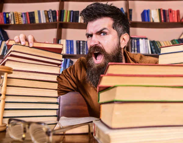 Lehrer oder Schüler mit Bart sitzen mit Brille am Tisch, defokussiert. Mann mit brüllendem Gesicht zwischen Bücherstapeln, während er in der Bibliothek studiert, Bücherregale im Hintergrund. Wahnsinniges Wissenschaftlerkonzept — Stockfoto