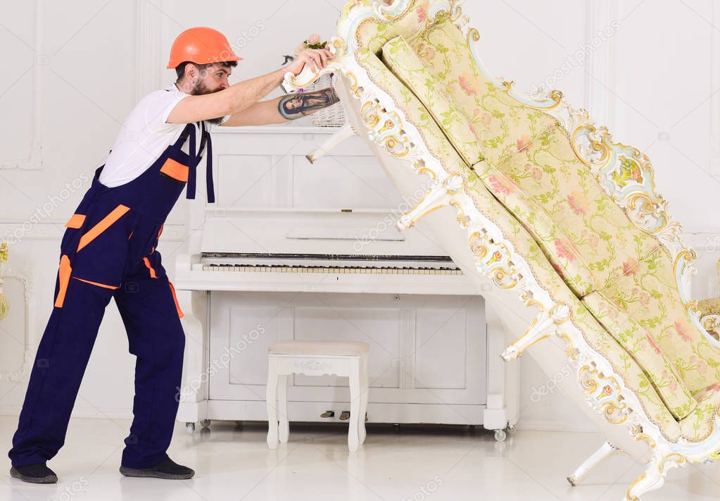 Delivery service concept. Courier delivers furniture in case of move out, relocation. Man with beard, worker in overalls and helmet lifts up sofa, white background. Loader moves sofa, couch