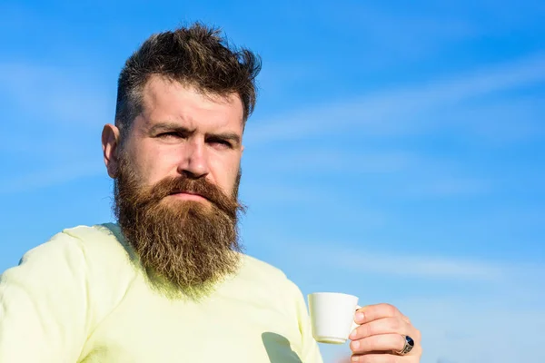 Man with long beard enjoy coffee. Coffee gourmet concept. Bearded man with espresso mug, drinks coffee. Man with beard and mustache on strict face drinks coffee, blue sky background, defocused