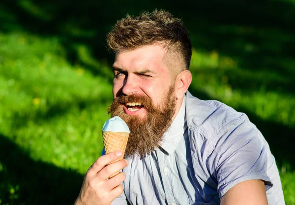 Concept de tentation. Homme barbu cône de crème glacée. L'homme à longue barbe mange de la crème glacée, tout en s'asseyant sur l'herbe. Homme avec barbe et moustache sur le visage clignant profiter de crème glacée, herbe sur fond déconcentré — Photo
