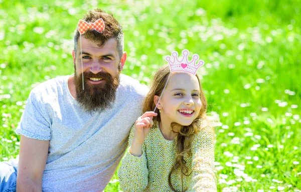 Dad and daughter sits on grass at grassplot, green background. Child and father posing with crown and bow photo booth attributes. Fatherhood concept. Family spend leisure outdoors, play girlish games — Stock Photo, Image
