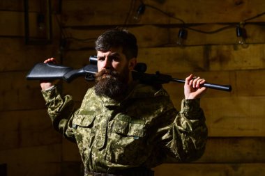 Hunter, brutal hipster on strict face with gun ready for hunting. Man, gamekeeper with beard wears camouflage clothing, carries rifle on shoulders, wooden interior background. Masculinity concept clipart