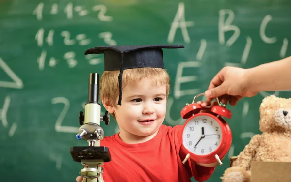 Kid boy in academic cap near clock in male hand, classroom, chalkboard on background. School break concept. Child on smiling face looks at alarm clock. Pupil waiting for school break