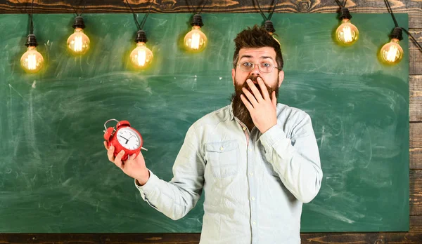 Forgot about time concept. Man with beard and mustache on shocked face in classroom. Bearded hipster holds clock, chalkboard on background, copy space. Teacher in eyeglasses holds alarm clock — Stock Photo, Image