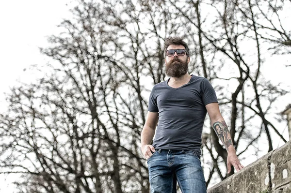 Homem hipster barbudo com braço tatuado em pé contra a antiga parede de pedra. Homem musculoso brutal vestindo óculos de sol na moda em cena urbana, moda e estilo — Fotografia de Stock