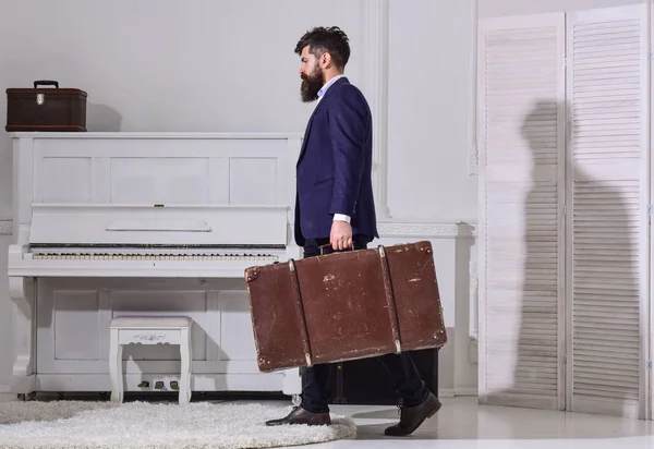 Macho con estilo en paseos estrictos por la cara y lleva una gran maleta vintage, vista lateral. Equipaje y concepto de viaje. Hombre, viajero con barba y bigote con equipaje, fondo interior blanco de lujo — Foto de Stock