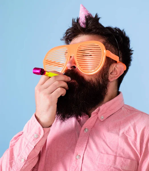 Man with beard on calm face holds party horn, light blue background. Guy in party hat with holiday attributes celebrates. Hipster in giant glasses blows into party horn. Surprise concept — Stock Photo, Image