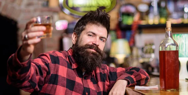 Guy spend leisure in bar, defocused background. Hipster with beard holds alcoholic beverage. Cheers concept. Man drinks whiskey or cognac. Man on relaxed face sits near bar counter, raising up glass