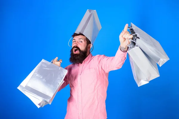 Hipster mit fröhlichem Gesicht und Tasche auf dem Kopf sind süchtig nach Shopaholic. Mann mit Bart und Schnurrbart trägt Einkaufstaschen, blauer Hintergrund. Einkaufskonzept. Mann kauft in der Verkaufssaison mit Rabatten ein — Stockfoto