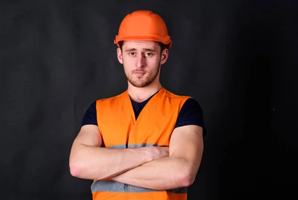 Man in helmet, hard hat hold arms crossed on chest, black background. Worker, contractor, builder on strict face with muscular biceps. Strong builder concept. Builder in helmet posing, copy space