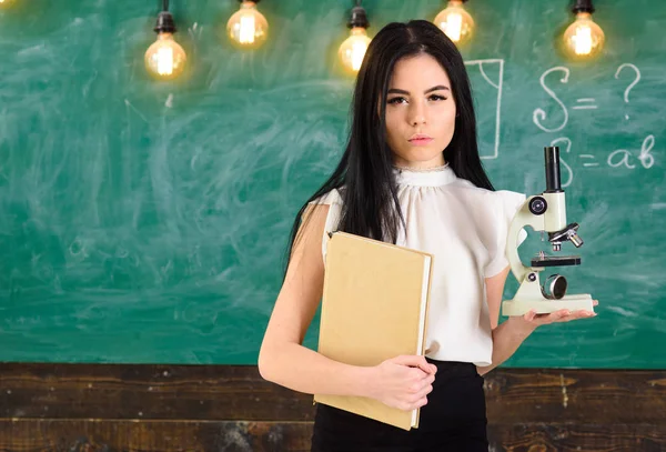 Leraar biologie houdt boek en Microscoop. Lady in formele slijtage op kalme gezicht in de klas. Biologie concept. Lady wetenschapper houdt boek en Microscoop, schoolbord op achtergrond, kopie ruimte — Stockfoto