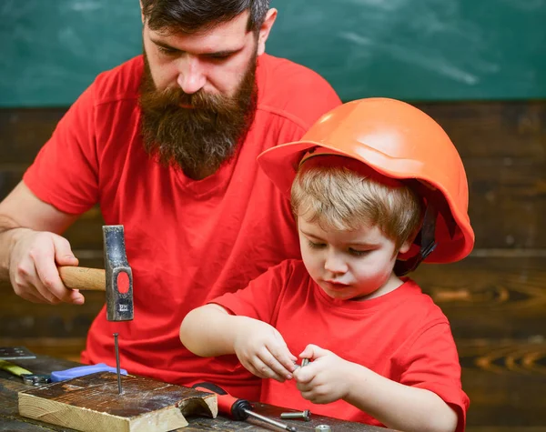 ひげ第とハンマーを使用少し息子を教育と親の父。少年は、保護用のヘルメットをパパと一緒に第槌で打つことの学習で忙しい子。ワーク ショップと便利屋の概念 — ストック写真