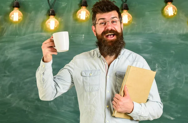 Homem com barba no rosto feliz na sala de aula.Professor em óculos detém livro e caneca de café ou chá. Conceito de pausa para café. Cientista detém livro e caneca de café, quadro no fundo, espaço de cópia — Fotografia de Stock