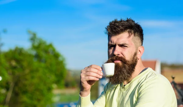 Homem barbudo com caneca de café, bebe café. Conceito de pausa para café. Homem com barba longa parece rigoroso e sério. Homem com barba e bigode no rosto rigoroso bebe café, fundo urbano, desfocado — Fotografia de Stock