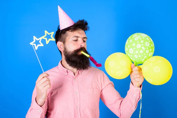 Hipster mit sternförmiger Brille bläst ins Partyhorn. Mann mit Bart auf ruhigem Gesicht hält Luftballons, blauer Hintergrund. Überraschungskonzept. Mann im Partyhut mit Urlaubsattribute feiert — Stockfoto