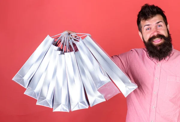 Hombre con barba y bigote sostiene bolsas de compras, fondo rojo. Hipster en la cara feliz es adicto a las compras o adicto a las compras. Concepto de compras. Guy de compras en temporada de ventas con descuentos —  Fotos de Stock