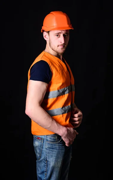 Concepto constructor atractivo. Constructor en casco posando con confianza. Hombre con casco, sombrero duro lleva chaleco naranja, fondo negro. Trabajador, contratista, constructor de cara seria con bíceps musculares — Foto de Stock