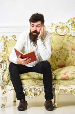 Maço sıkılmış yüz kitap okuma. Bilimsel literatür kavramı. Sakal ve bıyık oturur barok tarzı kanepe, tutar kitap, beyaz duvar arka plan üzerinde olan adam. Adam eski sıkıcı kitap okumak