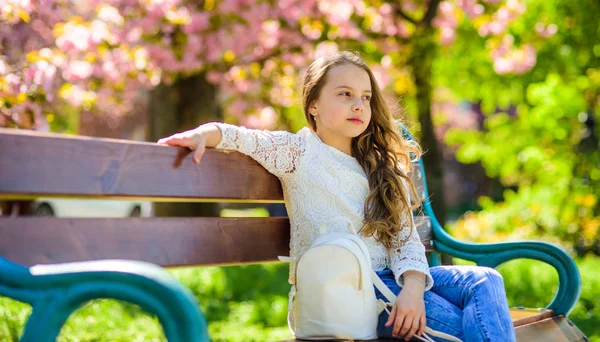 Chica en la cara sonriente se sienta en el banco, sakura en el fondo, desenfocado. Chica relajante mientras camina en el parque cerca de flor de cerezo. Lindo niño con mochila disfrutar de soleado día de primavera. concepto de accesorios de moda — Foto de Stock