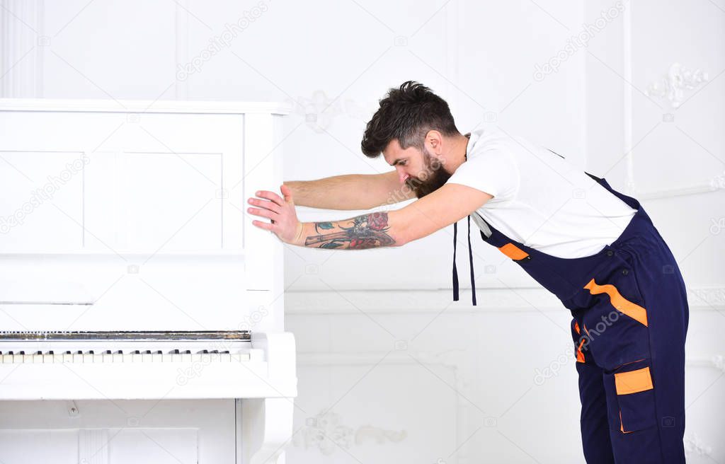 Man with beard and mustache, worker in overalls pushes piano, white background. Delivery service concept. Loader moves piano instrument. Courier delivers furniture in case of move out, relocation