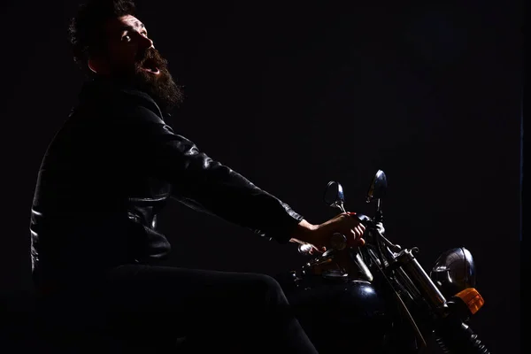 Homem com barba, motociclista de jaqueta de couro sentado em moto no escuro, fundo preto. Conceito de lazer para motociclistas. Macho, motociclista brutal em couro jaqueta equitação motocicleta à noite, espaço cópia — Fotografia de Stock