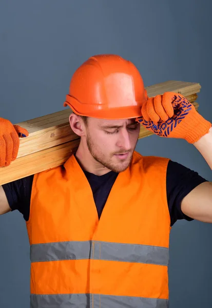 Carpenter, woodworker, strong builder on busy face carries wooden beam on shoulder. Safety and protection concept. Man in protective gloves holds visor of protective hard hat, grey background — Stock Photo, Image