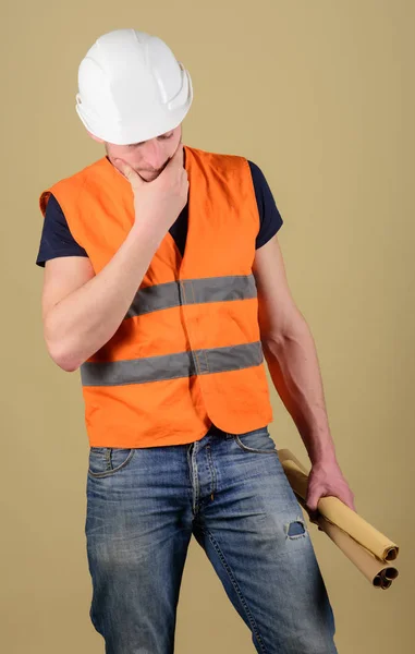 Ingegnere, architetto, operaio, costruttore sul viso premuroso tiene in mano vecchi progetti. Uomo, caposquadra in casco, cappello rigido ha dubbi sul piano edilizio, sfondo ocra. Concetto di ingegneria — Foto Stock