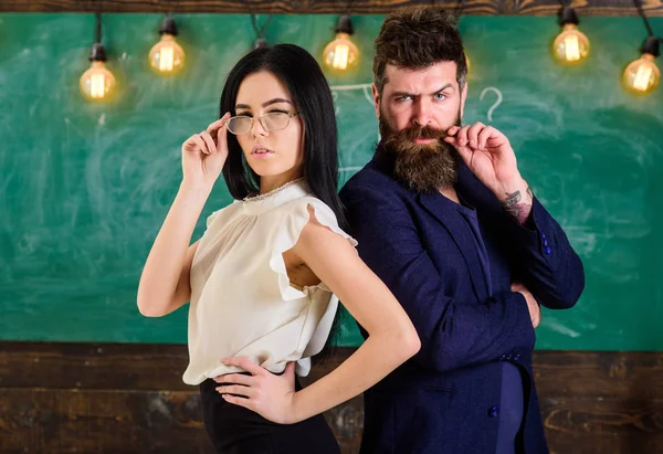 El hombre con barba y el maestro con anteojos se paran de espaldas, pizarra en el fondo. Dama e hipster trabajando juntos en la escuela. El maestro y el maestro parecen confiados. Concepto de trabajo en equipo — Foto de Stock